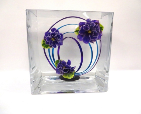 Underwater design using a glass cube with three double blue violet flowers and loops of purple and blue wire