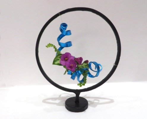 A miniature arrangement of African violet blossoms and blue curling wire suspended in a wrought iron stand